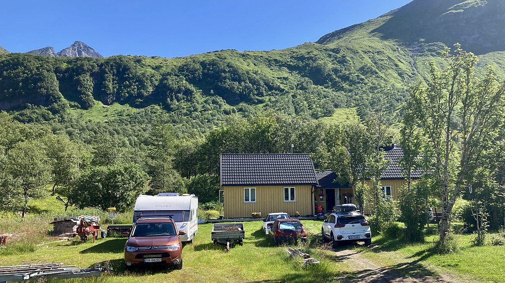 Stien til Strandtinden Store starter rett bak det gule huset Vassveien 66. Stien går opp skråningen rett bak huset. Store Strandtinden rager opp i bakgrunnen til venstre i bildet