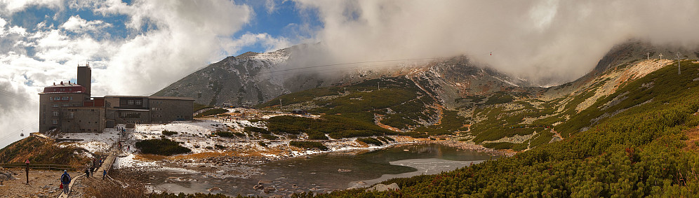 Ved Skalnate pleso med taubanestasjonen til venstre. Ser man godt etter ser man vogna på vei oppover mot toppen som en rød liten prikk.