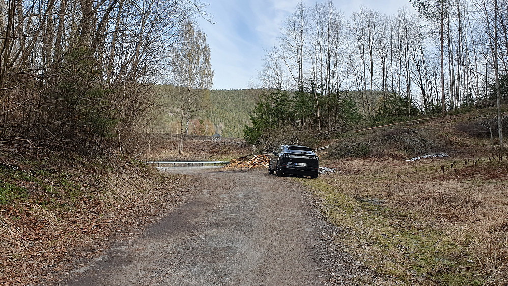 Parkering langs grusveien som går et lite stykke oppover Kyrkjeåsen (211). Noen meter forbi bilen går riksvei 36, og enda lengre bak ses spiret på Melum kirke. 